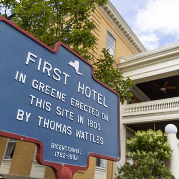 Blue NY State historic marker reading FIRST HOTEL IN GREENE ERECTED ON THIS SITE IN 1803 BY THOMAS WATTLES with hotel porch and columns behind